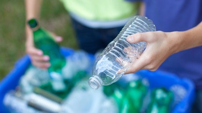 Caucasian boy and girl putting clear and green bottles and metal cans in recycling blue bin outside in yard